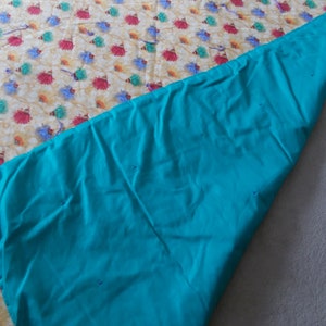 May include: A turquoise blue fabric backing is folded over a cream-colored fabric with a colorful pattern of flowers. The fabric is pinned together.