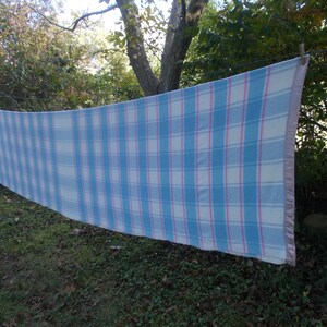 May include: A vintage blue and white plaid blanket with pink stripes is hanging on a clothesline outdoors. The blanket has a fringed edge and is made of a soft material. The blanket is a classic design, perfect for a picnic or adding a touch of nostalgia to a home.