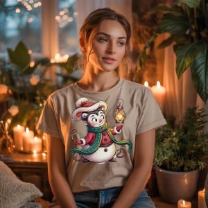 May include: A beige t-shirt featuring a cartoon snowman-mouse character wearing a red coat, green scarf, and a hat. The character is holding a lantern. The shirt is a short-sleeved crew neck.