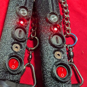 May include: Two black, textured devices with red illuminated buttons and keyhole-shaped openings. Each device is connected by a silver chain and a swivel clasp. The background is a solid red.