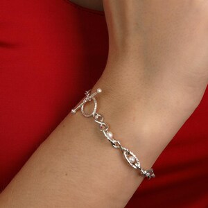 May include: A silver bracelet with a toggle clasp. The bracelet features a series of oval links, with small, round embellishments. The bracelet is worn on a wrist, against a red background.