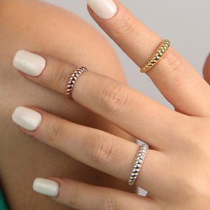 Könnte beinhalten: Vier Perlenringe in Gold, Roségold und Silber werden auf Fingern präsentiert. Die Ringe haben ein strukturiertes, gepunktetes Design. Die Person trägt ein türkisfarbenes Oberteil und hat weißen Nagellack.