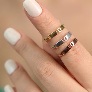 Könnte beinhalten: Nahaufnahme von drei minimalistischen Ringen in Gold, Silber und Roségold. Die Ringe sind dünne Bänder mit einem rechteckigen Design. Die Ringe werden an einem Finger vor einem verschwommenen Hintergrund getragen.