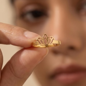 May include: A gold-colored ring featuring a lotus flower design. The ring is held up close to the camera, showcasing the intricate details of the floral pattern. The ring is a delicate piece of jewelry.