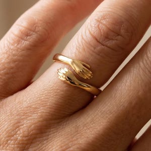 May include: A gold-colored ring featuring two hands embracing. The ring wraps around the finger with the hands clasped together. The ring is a simple, elegant design.