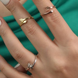 May include: Close-up of a hand wearing four arrow-shaped rings in gold, rose gold, and silver. The rings are on different fingers, with the arrowheads pointing in various directions. The background is a solid teal green.