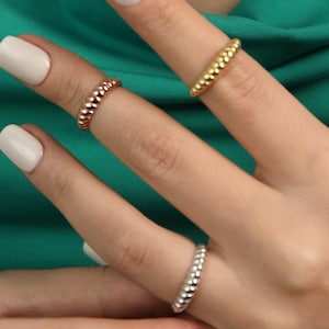 Op de afbeelding: Close-up van drie kralenringen in goud, roségoud en zilver. De ringen worden gedragen op vingers met witte nagellak, tegen een groene achtergrond. De ringen hebben een textuur, gestippeld ontwerp.