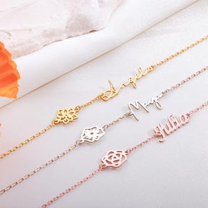 May include: Three delicate gold and rose gold bracelets. Each bracelet features a name in cursive script: "Angela", "Maya", and "Julia". Each bracelet has a floral charm. The bracelets are on a white surface.