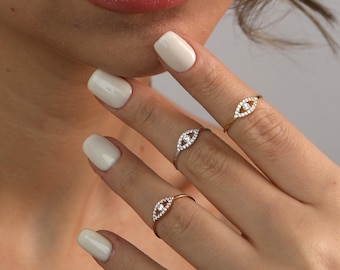 May include: Three delicate eye-shaped rings, each featuring a central clear stone surrounded by smaller, sparkling accents. The rings are displayed on a hand with white nail polish, showcasing the jewelry's design and detail. One ring is gold, one is silver, and one is rose gold.