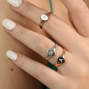 Könnte beinhalten: Drei Siegelringe mit eingravierten Initialen. Ein Ring ist roségoldfarben mit weißer Oberfläche und kleinem Herz. Der zweite Ring ist silberfarben mit dem Buchstaben "M". Der dritte Ring ist goldfarben mit dem Buchstaben "A".