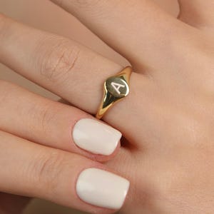 Könnte beinhalten: Ein goldener Siegelring mit dem Buchstaben "A" auf der ovalen Oberfläche. Der Ring wird an einem Finger getragen und zeigt seine polierte, reflektierende Oberfläche. Das Design des Rings ist schlicht und elegant.