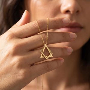 May include: A gold necklace with a geometric pendant composed of two overlapping diamond shapes. The pendant is suspended from a fine chain. The necklace is held up by a hand, highlighting the design and colour of the jewellery.