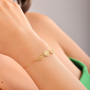 Puede incluir: Una delicada pulsera dorada con tres dijes: un ala, una flor y un pequeño pájaro. La pulsera se lleva en un brazo, con los dijes colocados en la muñeca. El fondo está borroso, resaltando la joyería.