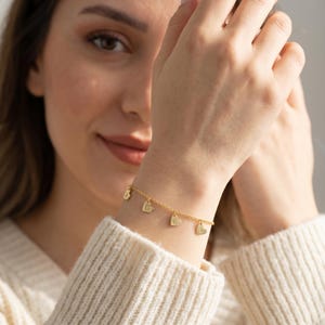 Könnte beinhalten: Ein goldfarbenes Armband mit herzförmigen Anhängern, die jeweils einen Buchstaben tragen. Das Armband wird am Handgelenk getragen, und die Anhänger hängen frei. Es ist ein zarter Schmuck.