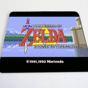 May include: A black mouse mat featuring a pixelated image of the video game "The Legend of Zelda: A Link to the Past". The image shows Link's sword, the game's title, and a background of a lake and mountains. The text "©1991,1992 Nintendo" is at the bottom of the image.