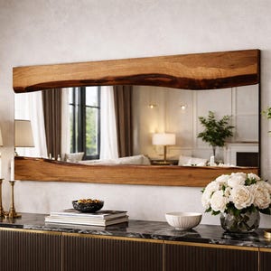 May include: A rectangular mirror with a wooden frame is mounted above a dark brown cabinet. The mirror reflects a room with curtains and a sofa. A vase of white roses sits on the cabinet, along with a lamp and decorative items.