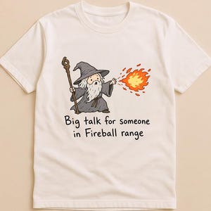 Könnte beinhalten: Ein cremefarbenes Kurzarm-T-Shirt mit einer Grafik eines Cartoon-Zauberers in grauem Gewand und Hut, der einen hölzernen Stab hält und einen leuchtend orangefarbenen Feuerball wirft. Darunter steht der Text: Big talk for someone in Fireball range.