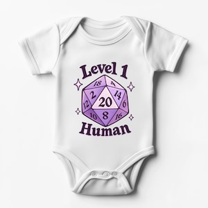 Könnte beinhalten: Ein weißer Babybody mit kurzen Ärmeln und einer violetten Grafik eines zwanzigseitigen Würfels in der Mitte. Der Text Level 1 steht über dem Würfel und Human darunter. Vier kleine vierzackige Sternsymbole umgeben die Würfelillustration.