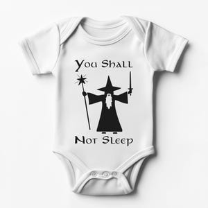 Può includere: Body da bambino bianco con grafica nera di un mago con bastone e spada. Il testo "You Shall Not Sleep" è sopra e sotto l'illustrazione. Il body ha maniche corte e chiusura con bottoni a pressione.