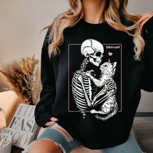 May include: Black sweatshirt featuring a white skeleton embracing a white cat. The design includes the text "United in spirit" and a pink heart. The sweatshirt is a casual garment.