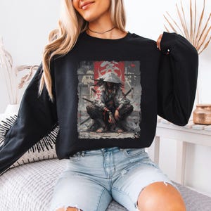 May include: Black sweatshirt featuring a detailed graphic of a samurai warrior in a crouching position, with swords and a red banner in the background. The warrior is wearing a hat and a mask. The sweatshirt is a long-sleeved crew neck.