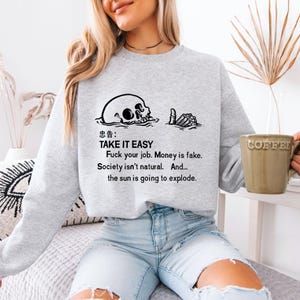 May include: A light gray sweatshirt with a black graphic of a skull and hand, with the text "TAKE IT EASY" and other phrases. A green coffee mug is held in the hand of a person.