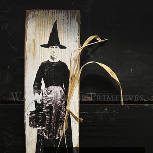 May include: A vintage-style wood sign with a black and white image of a woman in a witch's hat holding a basket. The sign has a white background with a distressed wood texture. The text on the sign reads "Elda Blackroot & Her Cat - Onyx 11"