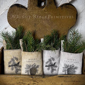 May include: Three small burlap pouches with a rustic, vintage look, each decorated with a black and white illustration of a bird on a branch, a pine cone, and the text "Merry Christmas". The pouches are displayed on a wooden shelf with a rustic, wooden heart-shaped sign above them that reads "Walnut Ridge Primitives".