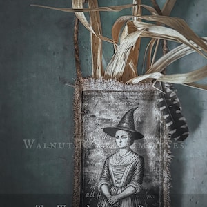 Op de afbeelding: Een rechthoekig jute zakje met gerafelde randen en een zwart-witte illustratie van een persoon met een puntmuts. Het zakje bevat gedroogde maïsbladeren en een gestreepte veer. Tekst: WALNUT RIDGE PRIMITIVES, THE WITCH'S HARVEST POCKET, 3 PRINTABLE GRAPHICS INCLUDED.