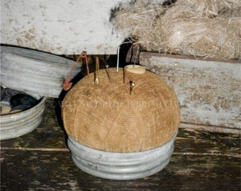 May include: A brown fabric pincushion with a metal base and several pins sticking out. The pincushion is sitting on a wooden surface. The text "The Farmer's Wife Pincushion" is visible at the top of the image.
