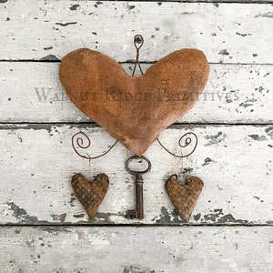 Könnte beinhalten: Ein braunes Stoffherz mit einem Schlüssel und zwei kleineren Herzen, die daran hängen. Die Herzen sind an einem Draht befestigt, der die Form einer Spirale hat. Der Hintergrund ist eine weiße Holzoberfläche.