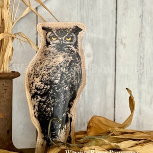May include: A black and white paper owl with yellow eyes. The owl is standing on a branch and has a rustic, aged look. The text "Walnut Ridge Primitives Aged Paper Owl - 10" is printed below the owl.