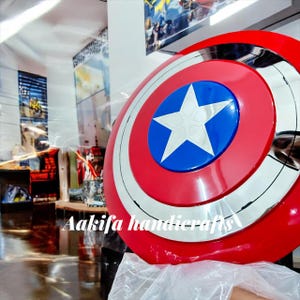 Peut inclure: Bouclier rond aux couleurs de Captain America : rouge, blanc et bleu, avec une &eacute;toile blanche sur un centre bleu. Bordure argent&eacute;e. Il s'agit probablement d'un accessoire ou d'un objet de collection.