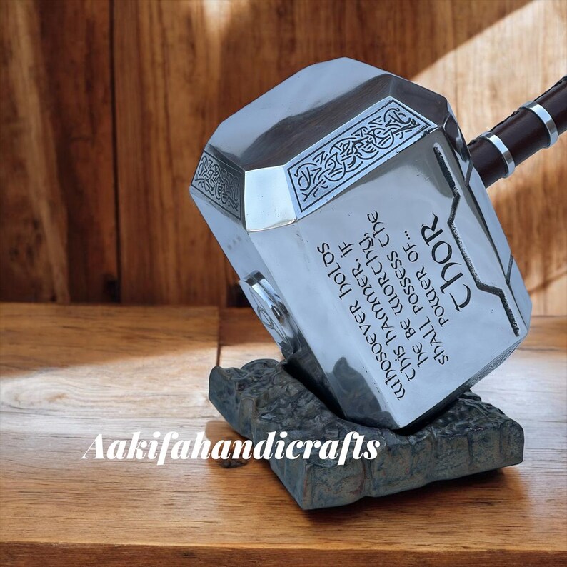 May include: A silver-coloured Thor's hammer replica with engraved text and a brown handle, resting on a grey stone base. The hammer features the inscription "WHOSOEVER HOLDS THIS HAMMER, IF HE BE WORTHY, SHALL POSSESS THE POWER OF... THOR."