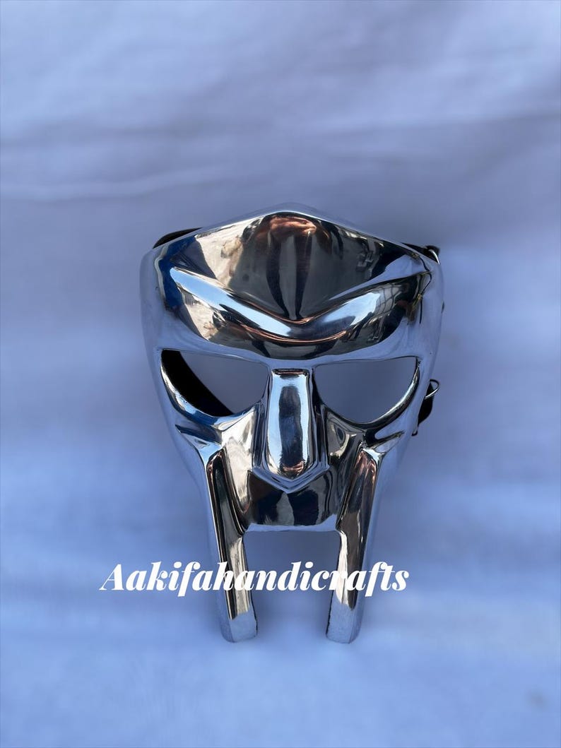 May include: A shiny silver mask with a unique, geometric design. The mask has a highly reflective surface and a stylized, tooth-like lower section. The text "Aakifahandicrafts" is printed on the bottom of the mask.