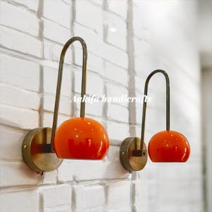 Peut inclure: Deux appliques murales avec des abat-jours globes oranges et des bras courbés en laiton. Elles sont montées sur des bases rondes en laiton contre un mur de briques blanches. Le texte "Aakifa handicrafts" est visible.