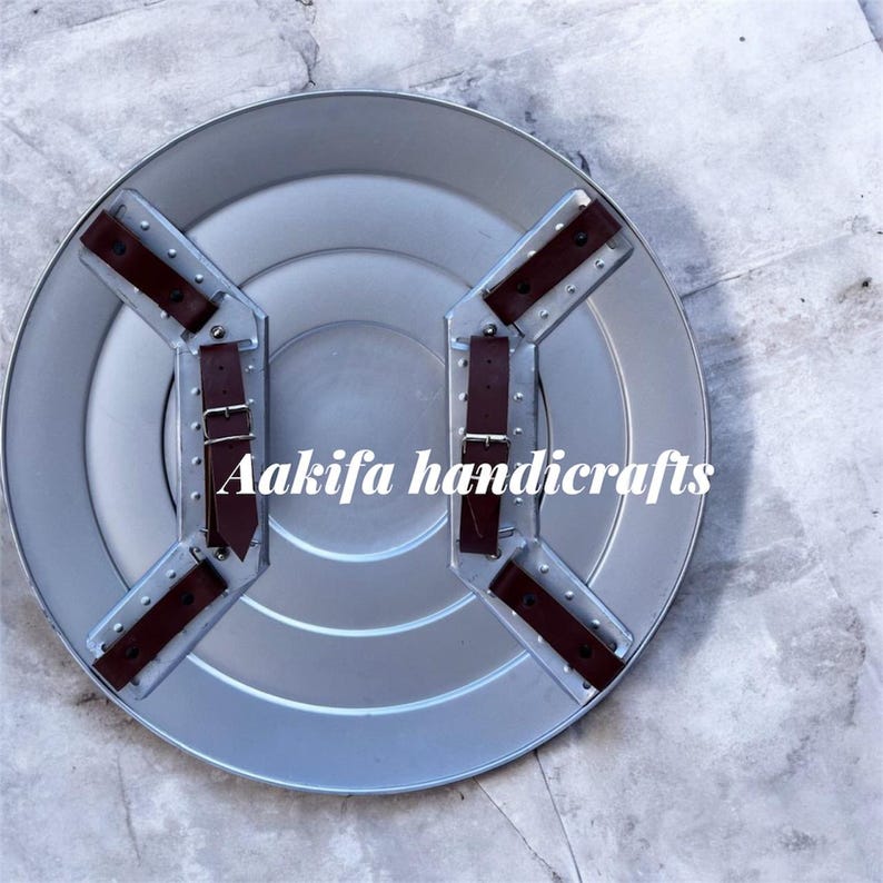 May include: A silver shield with brown leather straps and buckles. The shield has a circular design with a ridged centre. The text "Aakifa handicrafts" is printed on the shield.