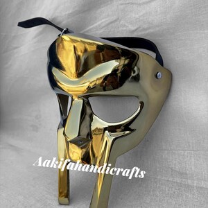 May include: A gold-coloured mask with a reflective surface, featuring a stylized design. The mask has a black strap and the text "Aakifahandicrafts" is printed on the front. The mask is likely for decorative or costume purposes.