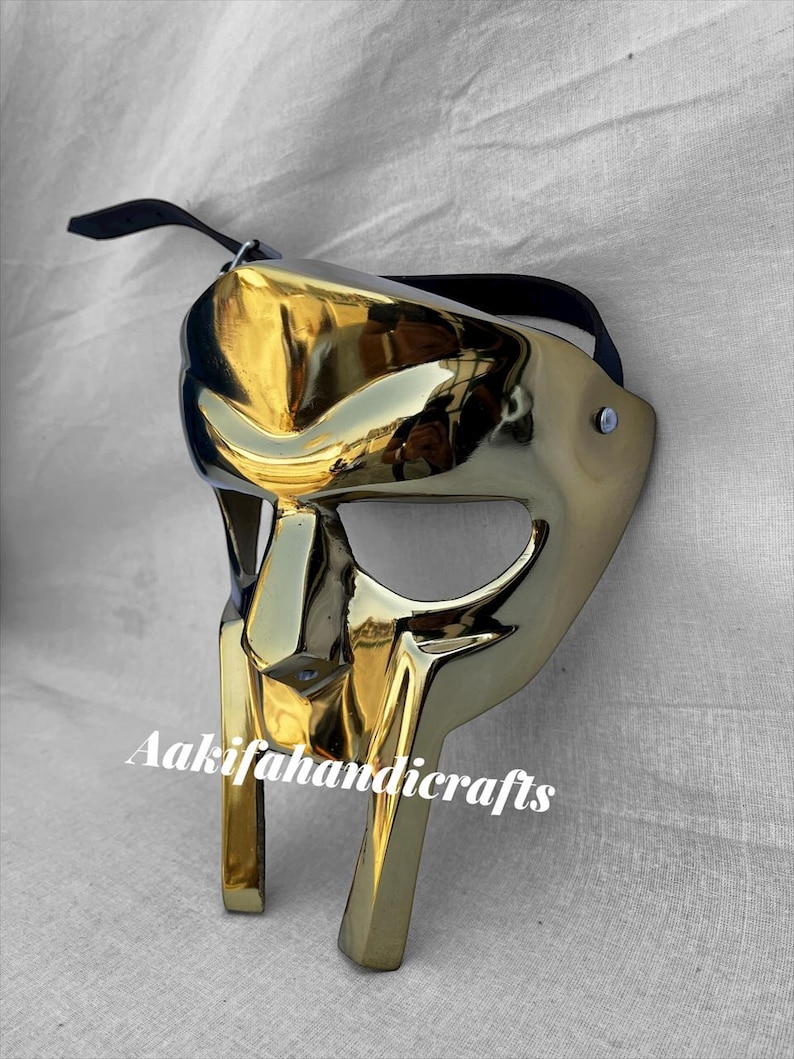 May include: A gold-coloured mask with a reflective surface, featuring a stylized design. The mask has a black strap and the text "Aakifahandicrafts" is printed on the front. The mask is likely for decorative or costume purposes.