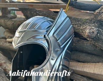 May include: A silver-colored helmet with a detailed design, featuring a curved visor and side panels. The helmet is placed on a wooden surface. The text "Aakifahandicrafts" is visible on the helmet.