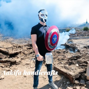 May include: A person wearing a silver mask, black shirt, and blue jeans holds a red, white, and blue shield with a star and a silver hammer. The image has the text "Aakifa handicrafts".