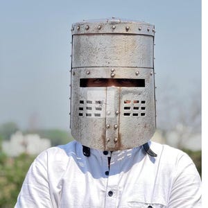 Può includere: Un elmo in metallo argentato, rivettato, con aperture rettangolari per occhi e bocca. L'elmo è di forma cilindrica, possibilmente storico o da costume. La persona indossa una camicia bianca.