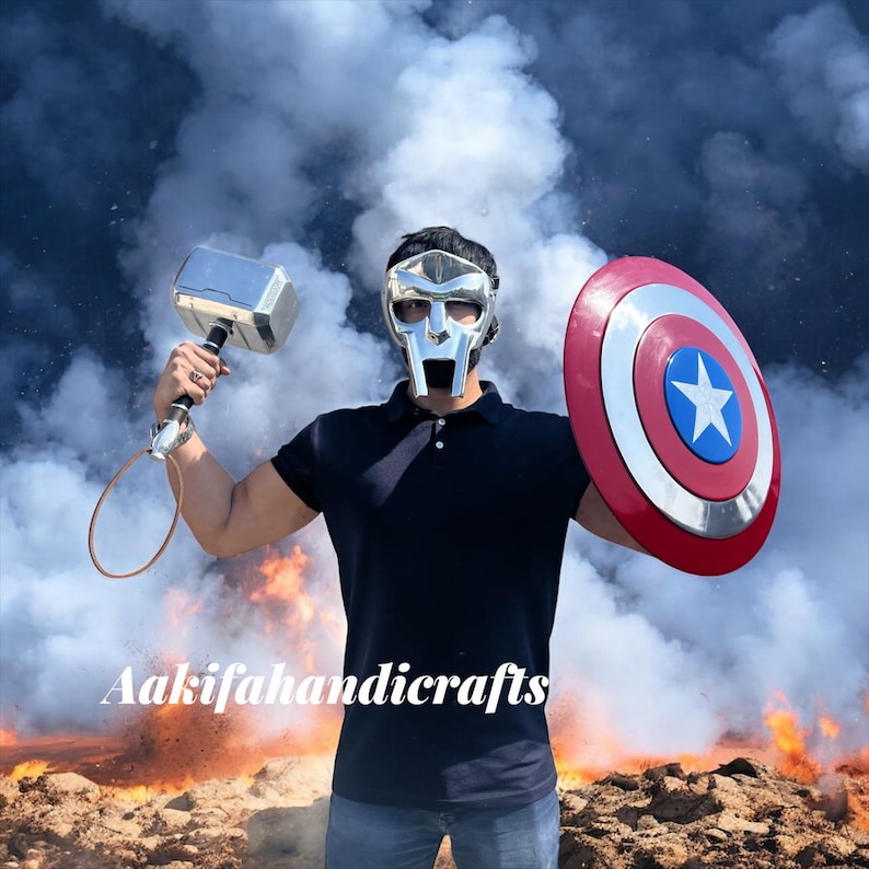 May include: A silver metal hammer and a red, white, and blue shield with a star are held up. The shield is round. The person is wearing a silver mask and a black polo shirt. The background has smoke and fire.