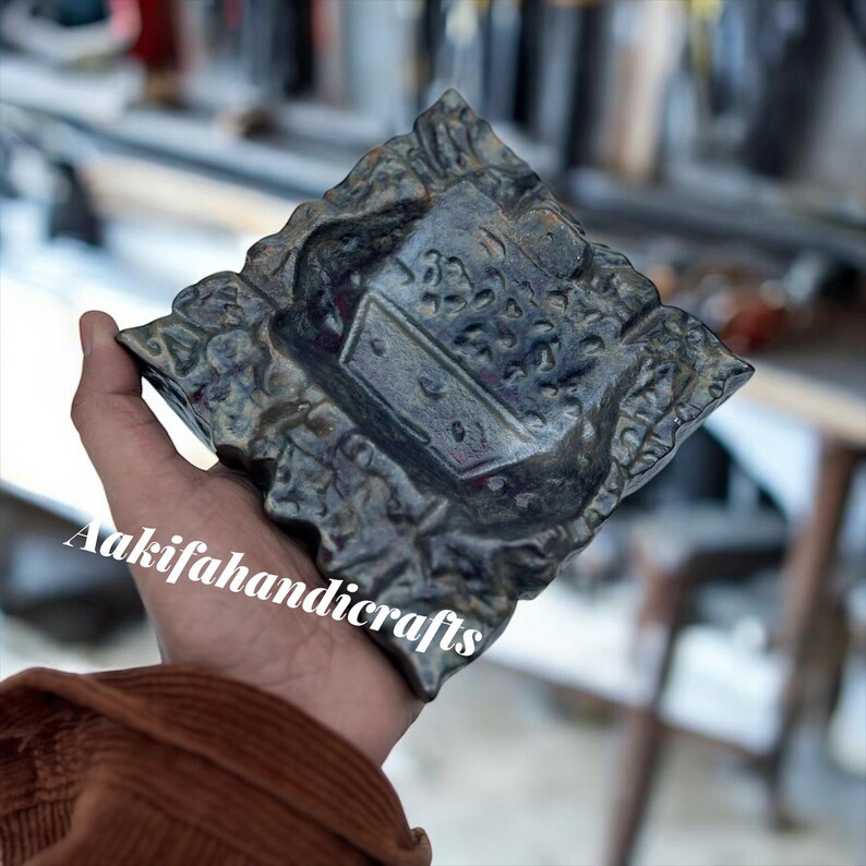 May include: A dark grey, square ashtray with a textured, floral-like design around the edges. The centre features a rectangular area with small holes. The text "Aakifahandicrafts" is visible on the bottom left.