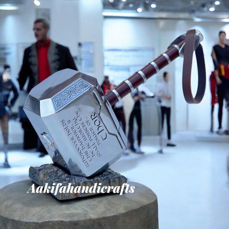 May include: A silver Thor's hammer replica with a brown handle and a leather strap. The hammer is engraved with text and rests on a stone base. The text on the hammer reads "Whoever holds this hammer, if he be worthy, shall possess the power of... Thor."