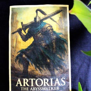 Può includere: Una stampa rettangolare con un guerriero in armatura scura, che tiene una grande spada. L'immagine ha uno schema di colori giallo e blu, con il testo "ARTORIAS THE ABYSSWALKER" in basso. La stampa è su una superficie nera.