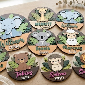 May include: A collection of circular wooden plaques featuring layered 3D animal cutouts including an elephant, giraffe, koala, sheep, rabbit, bear, hedgehog, and lion, each set against green leaves with personalised names printed on the base.