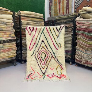 May include: Cream-colored rug with geometric designs in pink, green, yellow, and black. The rug features a diamond pattern in the center and colorful lines. The rug is displayed in a shop with other rugs and textiles.