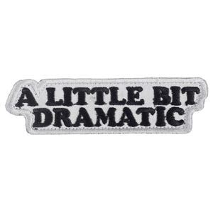 May include: A rectangular patch with the phrase "A LITTLE BIT DRAMATIC" embroidered in black on a white background. The patch has a white border with a stitched edge.