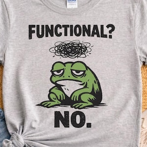 May include: A light gray t-shirt featuring a green frog with a grumpy expression. The shirt has the text "FUNCTIONAL?" above the frog and "NO." below it, all in black. The shirt is made of a soft material.